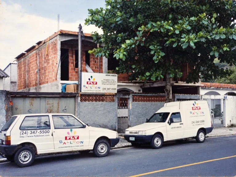primeira sede da Fly Balloon em 1998, com sobrado em construção, placa da empresa e dois veículos com a marca Fly Balloon estacionados na frente
